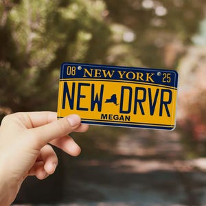 Puede incluir: Una placa de matr&iacute;cula amarilla y azul de Nueva York con el texto "NEW DRVR" y el nombre "Megan". La placa tambi&eacute;n tiene el texto "NEW YORK" y los n&uacute;meros "08" y "25".