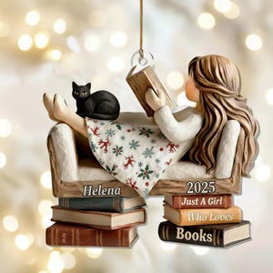 May include: A decorative ornament featuring a girl reading a book while reclining on a sofa with a black cat. The girl is wearing a white dress with a patterned skirt. The ornament includes the text "Helena" and "2025 Just a Girl Who Loves Books."