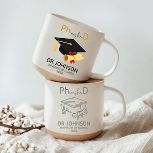 May include: Two stacked white ceramic mugs with a graduation theme. The top mug features a black graduation cap, a diploma, and the text "Phinishe D, DR. JOHNSON, UNIVERSITY OF FLORIDA 2026." The bottom mug has a graduation cap and diploma graphic.