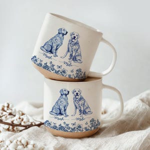 May include: Two stacked ceramic mugs with white backgrounds and brown bases. Each mug features two dog illustrations in blue, with floral and butterfly accents. The names "Mush" and "Milo" are written below the dogs.