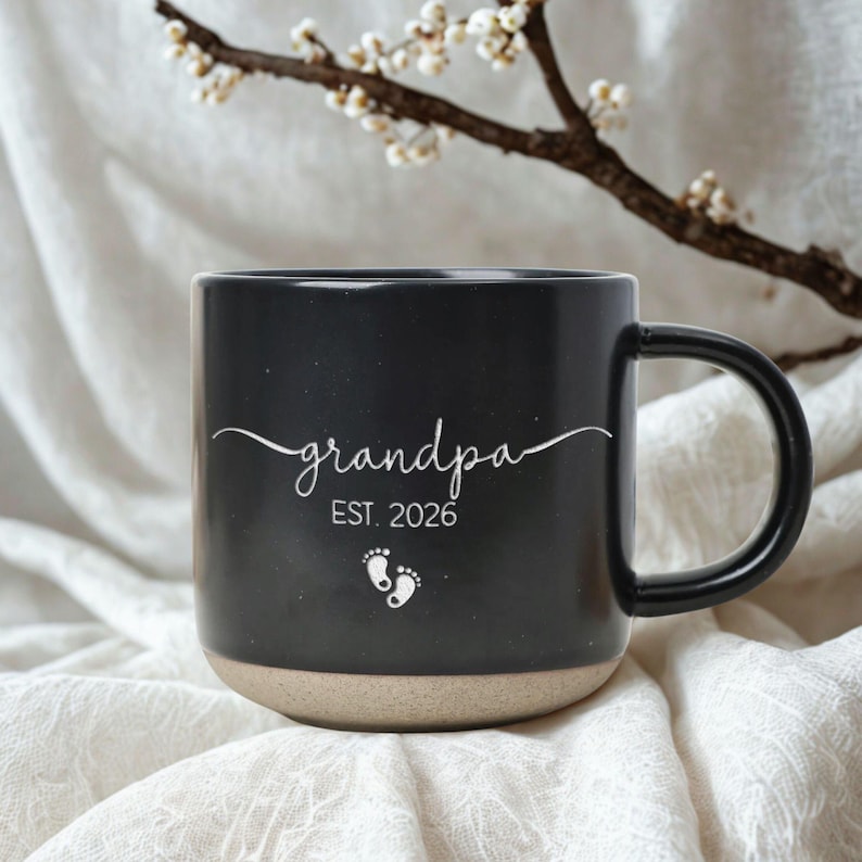 May include: Black ceramic mug with the word "grandpa" in white script, the year "EST. 2026", and two baby footprints. The bottom of the mug is a tan color. A black handle is on the right side.