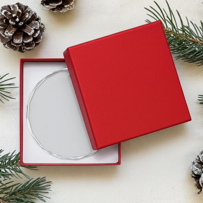 May include: A clear, circular ornament with a silver edge sits in a red gift box with a white interior. The box is partially open, revealing the ornament. Pinecones and evergreen sprigs decorate the background, suggesting a festive theme.