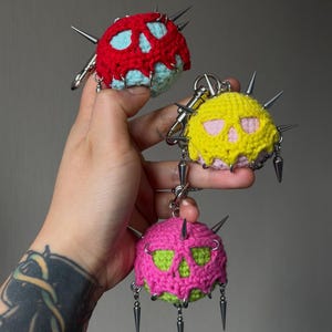 Puede incluir: Tres llaveros de ganchillo esféricos con recortes en forma de calavera. El superior es rojo con azul claro, el medio es amarillo con rosa y el inferior es rosa con verde lima. Cada pieza está adornada con púas de metal plateado.