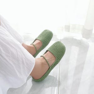 Könnte beinhalten: Ein Paar grüne Häkelslipper mit einem Riemen über dem Fuß. Die Slipper sind aus einem strukturierten Garn gefertigt und scheinen handgefertigt zu sein. Die Slipper werden von einer Person getragen, die ein weißes Kleid trägt.