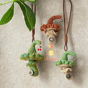 Pode incluir: Três pingentes de cogumelos de crochê feitos à mão em tons de verde, marrom e bege, cada um com um botão e um cordão de couro. Um pingente tem um pingente de cereja. Os pingentes são exibidos em um fundo neutro.