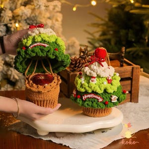 Könnte beinhalten: Zwei gehäkelte Weihnachtsbaumdekorationen. Eine ist ein Korb mit einem Apfel, die andere ein Baum mit roten Akzenten und dem Text "Happy Everyday". Beide sind grün, weiß und braun.