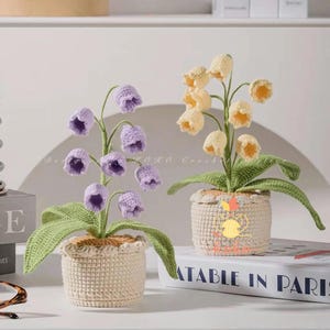 Może przedstawiać: Dwie szydełkowe kompozycje kwiatowe. Jedna ma fioletowe kwiaty w kształcie dzwonków, druga żółte kwiaty. Obie znajdują się w beżowych szydełkowych donicach z zielonymi liśćmi. Kompozycje stoją na białej powierzchni.