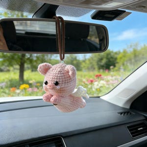 Puede incluir: Un adorno de coche de cerdo rosa tejido a mano, con pequeñas alas blancas, ojos negros y una lágrima. El cerdo está suspendido de un cordón marrón, colgando del espejo retrovisor de un coche. El fondo muestra un día soleado con un campo de flores.