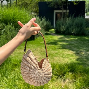May include: A beige, crocheted handbag with a unique, shell-like design. The bag has a brown leather strap and gold-colored hardware. The bag is held up by a person's hand in a grassy outdoor setting.