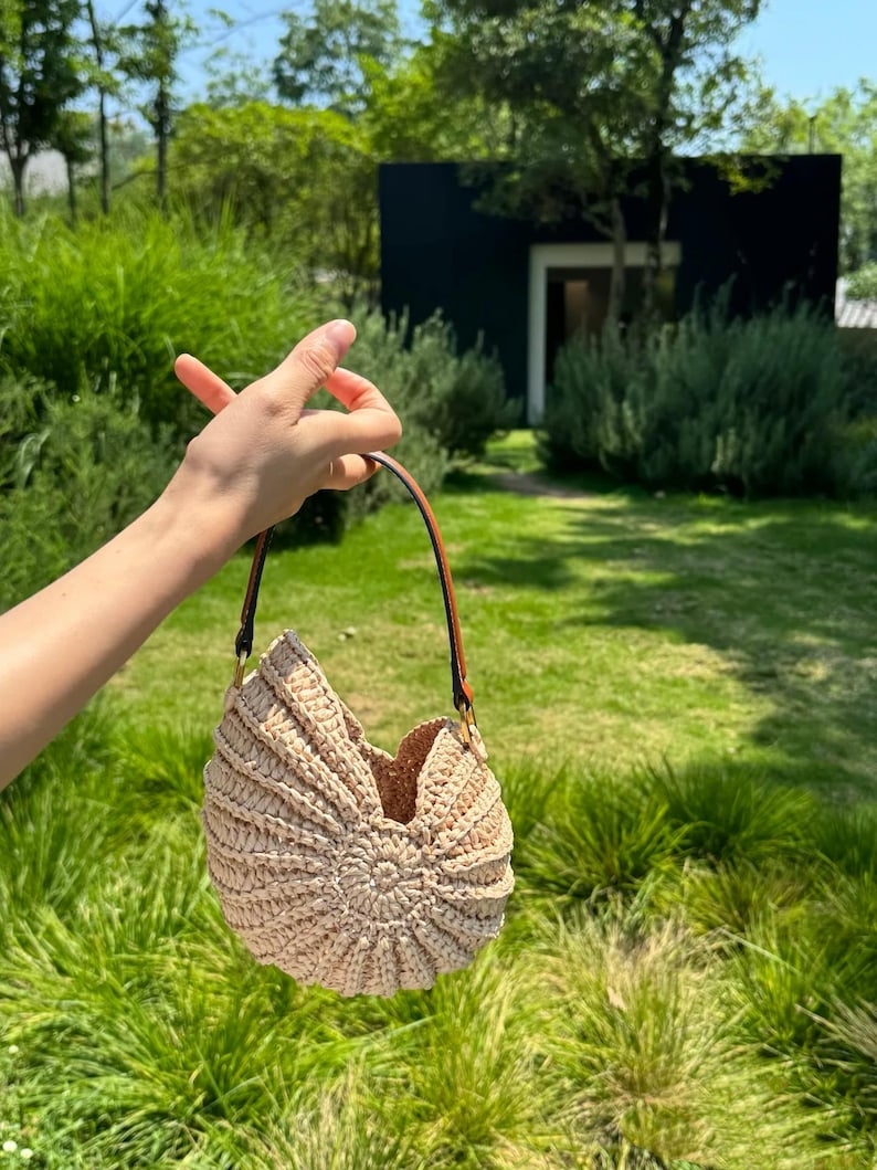 May include: A beige, crocheted handbag with a unique, shell-like design. The bag has a brown leather strap and gold-colored hardware. The bag is held up by a person's hand in a grassy outdoor setting.