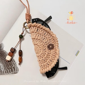 以下が含まれることがあります： 黒いサングラスを収納する、茶色のボタンが付いたベージュ色の織りサングラスケース。木製のビーズと緑色のビーズが付いた茶色の革紐に付いています。「koko」の文字が見えます。