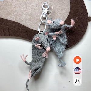 May include: A handmade crochet keyring featuring two grey rats with pink detailing. The rats have black whiskers and are attached to a silver-coloured keyring. The background is a light brown and white.