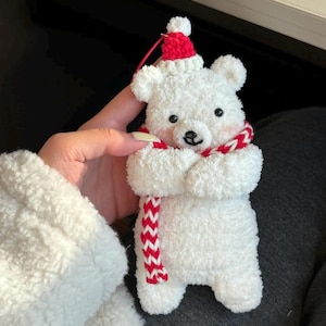 May include: A white plush polar bear ornament with a red and white scarf and a red hat. The bear has black eyes and a small black nose. The ornament is approximately 4 inches tall.