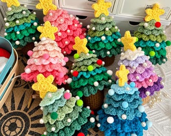 Op de afbeelding: Een verzameling kleine, kegelvormige gehaakte kerstbomen in diverse kleuren zoals groen, roze, blauw en paars. Elke boom is bekroond met een gele gehaakte ster en versierd met witte parelkralen en kleine kleurrijke pompons.