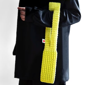 May include: A vibrant yellow crocheted tote bag with a textured, grid-like design. The bag features a long strap and a rectangular shape. The person in the image is wearing a black long-sleeved top. A small white tag with a red apple is visible.