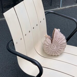 May include: A handmade, light brown crochet baby carriage sits on a cream-colored wooden bench with black metal arms. The carriage has a rounded shape and a textured surface, suggesting a soft, cozy design. The bench has a modern, minimalist style.