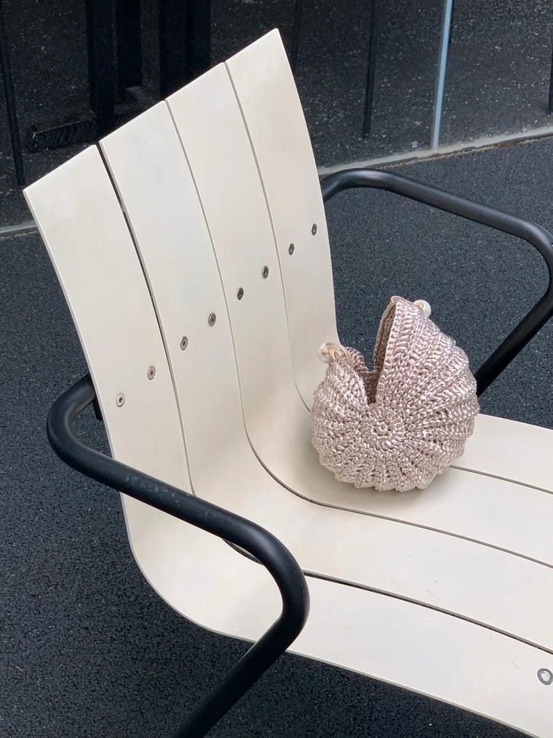 May include: A handmade, light brown crochet baby carriage sits on a cream-colored wooden bench with black metal arms. The carriage has a rounded shape and a textured surface, suggesting a soft, cozy design. The bench has a modern, minimalist style.
