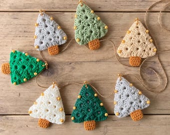 May include: A garland featuring seven crocheted Christmas tree ornaments in various colors including green, light green, white, cream, and light gray. Each tree has a brown crocheted trunk and small yellow beads attached to the edges.