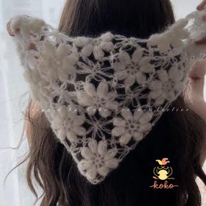 May include: A white, crocheted floral headscarf. The triangular scarf features large, three-dimensional flower designs. The text "Designed by Koko Crochet Atelier" is visible. The scarf is held up, showcasing the intricate details of the handmade piece.