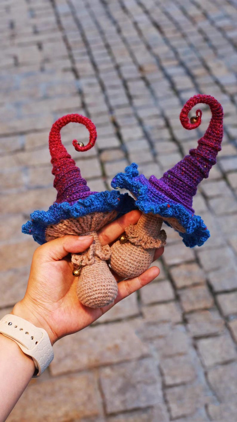 May include: Two handmade crochet mushroom hats with curled tips. The hats feature a color gradient from red to purple, blue frills, and a tan mushroom cap. Each hat has a small gold bell at the tip and base.