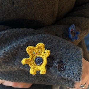 May include: A close-up of a gray wool coat sleeve with a yellow crocheted teddy bear applique and a blue crocheted flower applique. The teddy bear has a brown button in the center. The coat has dark brown buttons.