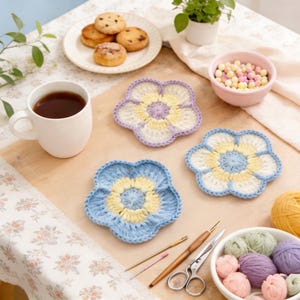 Könnte beinhalten: Handgefertigte gehäkelte Blumenuntersetzer in Blau, Gelb und Weiß. Die Untersetzer liegen auf einer Holzoberfläche, zusammen mit einer Tasse Tee, Keksen und Bastelzubehör. Die Szene vermittelt eine gemütliche, handwerkliche Ästhetik.