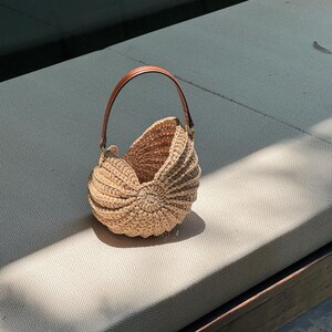 May include: A beige, nautilus-shaped crochet bag with a brown leather handle. The bag is made of a woven material and has a rounded shape with a spiral design on the front. The handle is attached to the bag with gold-colored hardware.
