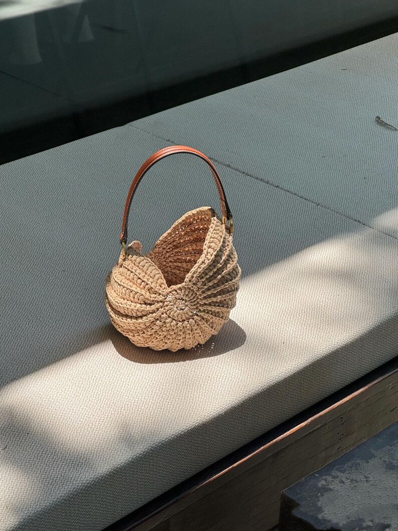 May include: A beige, nautilus-shaped crochet bag with a brown leather handle. The bag is made of a woven material and has a rounded shape with a spiral design on the front. The handle is attached to the bag with gold-colored hardware.