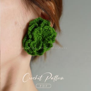 Op de afbeelding: Close-up van een felgroene, gehaakte oorbel in de vorm van een bloem. De oorbel wordt op een oor gedragen. De tekst "Crochet Pattern PDF" staat onderaan de afbeelding. De tekst "designed by The Bucket Lists" staat bovenaan.