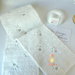 Puede incluir: Una bufanda blanca y esponjosa de ganchillo con recortes de huellas de pata. También se ven un ovillo de lana blanca y una pequeña tarjeta con texto escrito a mano. La bufanda está hecha de un material suave y texturizado.