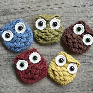 Puede incluir: Cinco objetos hechos a mano con forma de búho de ganchillo en varios colores: azul, verde, marrón, rojo y amarillo. Cada búho tiene ojos grandes y redondos con detalles blancos y negros. Los búhos están colocados sobre una superficie de madera.