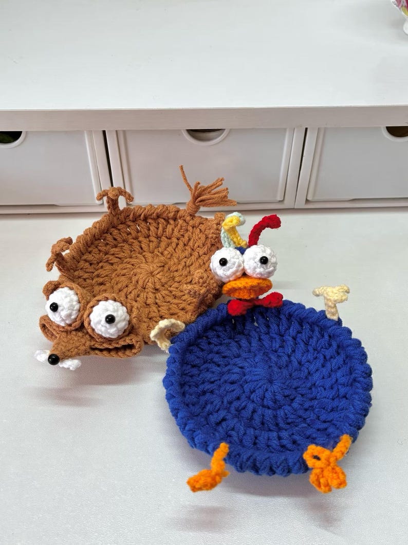May include: Two handmade crochet coasters. One is brown, resembling a cartoon animal with large white eyes and a snout. The other is blue, shaped like a bird with orange feet and a red beak. Both have white and black eyes.
