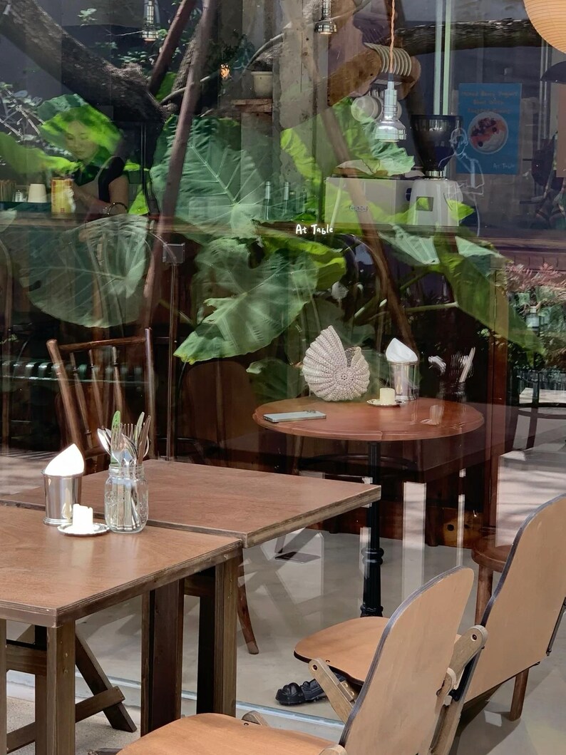 May include: A cafe interior with wooden tables and chairs. A round table has a decorative shell and a small plate. The text "At Table" is visible on the glass. Large green leaves are reflected in the glass.