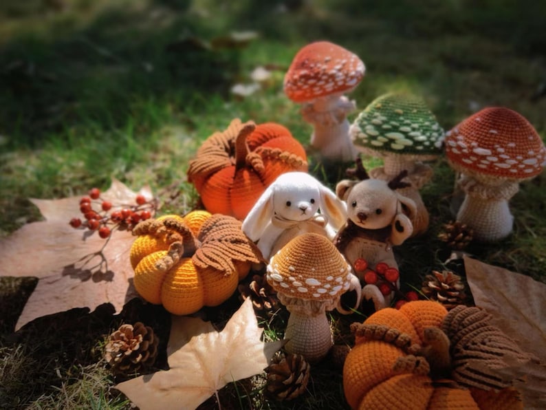 Puede incluir: Una colecci&oacute;n de art&iacute;culos de ganchillo hechos a mano, que incluyen calabazas, setas, un conejito blanco y un osito de peluche. Las calabazas son naranjas y amarillas, y las setas tienen sombreros rojos y verdes. La escena est&aacute; ambientada sobre un lecho de hojas y hierba.