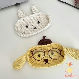 May include: Two handmade crochet trays, one white bunny-shaped and one yellow character-shaped with glasses. The bunny tray has black eyes and a stitched mouth. The yellow tray has brown details and holds a pair of round glasses.