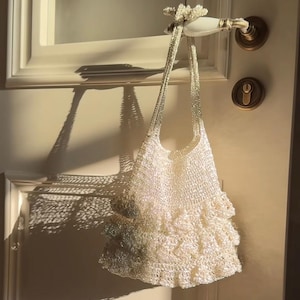 May include: A white, crocheted handbag with a ruffled bottom and beaded straps hangs from a door handle. The bag is made of a shimmering, light-colored material, and the door handle is gold.