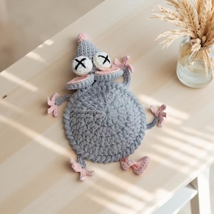 May include: A handmade crochet monster in gray, pink, and white. The monster has a round body, a pointed hat, and two eyes with black X's. The arms and feet are pink. The monster is lying on a light-colored wooden surface.