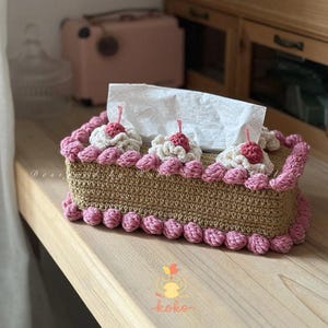 Può includere: Una copertura per scatola di fazzoletti fatta a mano con un design a forma di torta. La copertura è all'uncinetto in colore beige con bordi rosa e decorazioni di crema e ciliegie. La scatola di fazzoletti è su una superficie di legno.