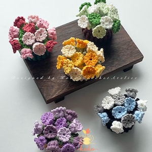 Op de afbeelding: Een verzameling handgemaakte gehaakte bloemstukken in verschillende kleuren, waaronder roze, rood, geel, wit, groen, blauw en paars. De bloemen zijn gerangschikt in kleine, ronde clusters, sommige op een houten dienblad. De tekst "Designed by Koko Crochet Atelier" is zichtbaar.