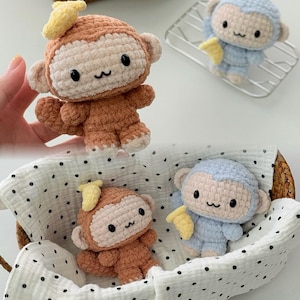 May include: Crocheted monkey toys in shades of brown and blue, each holding a yellow banana. The monkeys have black eyes and a smiling mouth. One monkey is held by a hand, while others are in a basket.