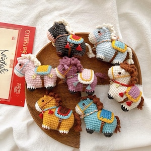 Könnte beinhalten: Eine Sammlung von sieben gehäkelten Pferdfiguren in verschiedenen Farben, darunter Rosa, Lila, Blau und Gelb. Jedes Pferd hat einen Sattel und eine Mähne, angeordnet auf einem Holztablett.