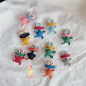 Könnte beinhalten: Eine Sammlung handgefertigter Häkelfiguren in Form von Fröschen. Jeder Frosch hat große Kulleraugen und einen breiten Mund, mit Farbvariationen in Rosa, Blau, Gelb, Grün, Rot, Orange und Grau. Die Frösche sind auf einer weißen, strukturierten Oberfläche angeordnet.