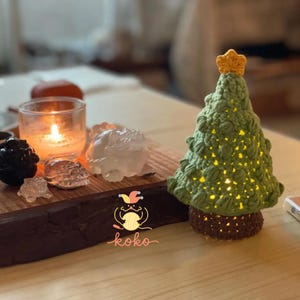 May include: A handcrafted, green crochet Christmas tree with a yellow star on top. The tree is illuminated from within, casting a warm glow. The base is brown. The tree is on a wooden surface with a lit candle and decorative crystals.
