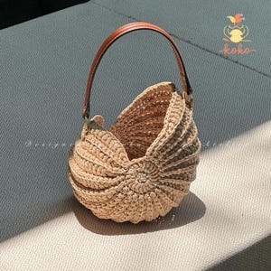 May include: A beige crochet handbag with a seashell design. The bag has a brown leather handle and a rounded shape. The bag is made of a thick, textured yarn and has a unique, handcrafted appearance. The bag is sitting on a grey surface.
