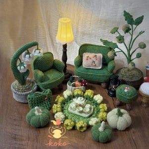 Op de afbeelding: Een verzameling handgemaakte gehaakte items, waaronder miniatuur groene fauteuils, een lamp, een plant, pompoenen en een theeservies. De items zijn gerangschikt op een houten oppervlak, met een zachte, neutrale achtergrond. Het thema is een gezellige, miniatuur woonkamer.
