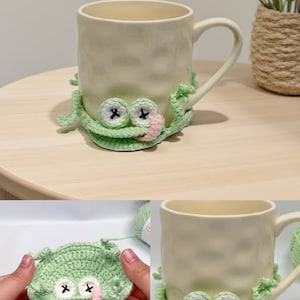 May include: A cream-colored ceramic mug adorned with a crocheted frog coaster. The frog coaster is light green with black cross-shaped eyes and a pink tongue. The mug is on a light brown surface.