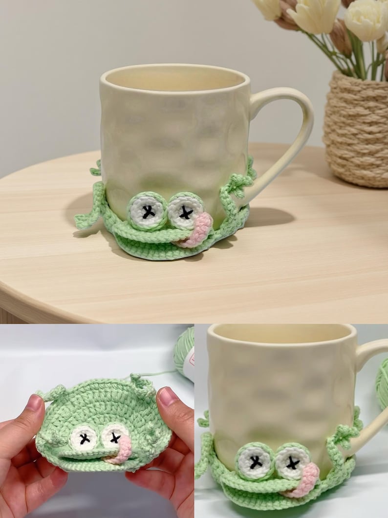 May include: A cream-colored ceramic mug adorned with a crocheted frog coaster. The frog coaster is light green with black cross-shaped eyes and a pink tongue. The mug is on a light brown surface.