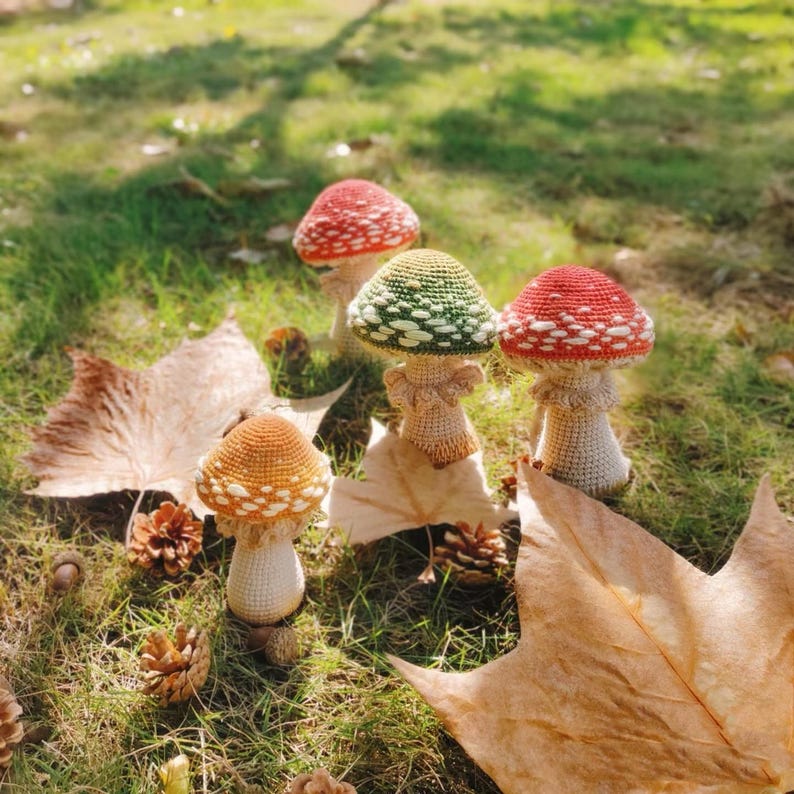 Puede incluir: Un conjunto de setas hechas a mano con ganchillo, en varios colores, incluyendo rojo, verde y naranja, con lunares blancos. Las setas est&aacute;n dispuestas sobre hierba verde, rodeadas de hojas ca&iacute;das y pi&ntilde;as, creando una escena oto&ntilde;al.