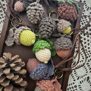 May include: A collection of handmade crochet acorn pendants in various colors, including brown, green, yellow, and blue. Each pendant is attached to a brown leather cord with a gold-colored ring. The pendants are displayed on a wooden tray.