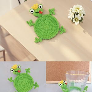 May include: A green crocheted frog coaster with a yellow crown and red tongue. The frog has large white-rimmed eyes and small green limbs. The coaster is round and sits on a light-colored wooden surface. A small bouquet of white flowers is also present.